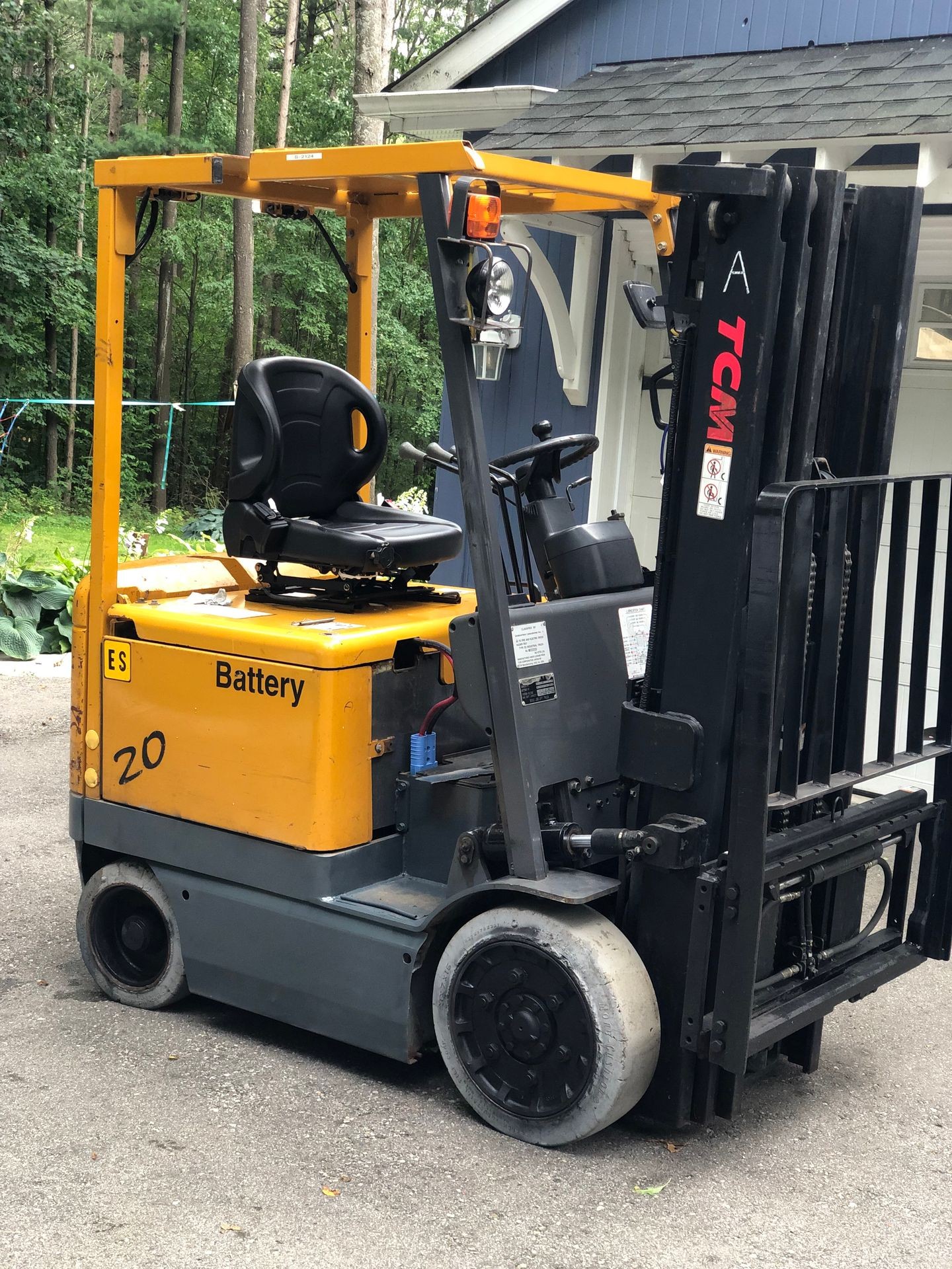 Yellow battery-operated forklift parked on a driveway with trees in the background.