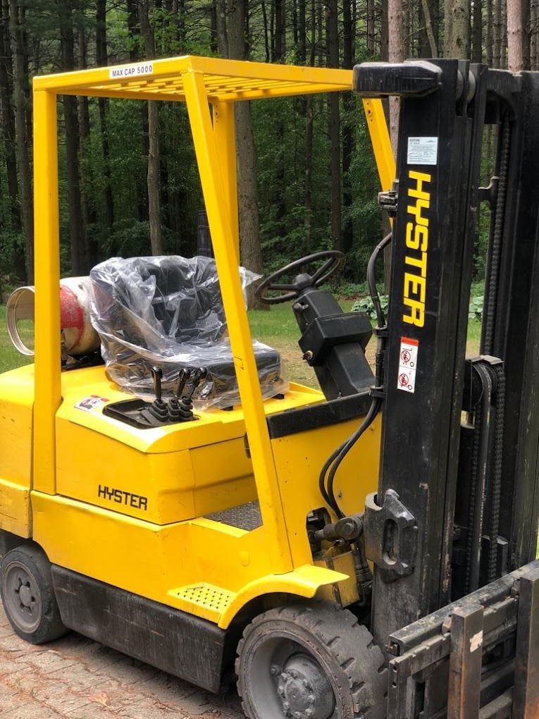 Yellow Hyster forklift parked on a wooded path, with a plastic-covered seat and lifting mechanism.